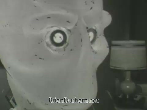 The still features a close-up of a stylized, expressionless face, possibly made from a plastic or similar material. It has large, round eyes that appear to be mechanical or cartoonish. The background includes a table lamp, contributing to a domestic ambiance. The image has a retro aesthetic typical of 1960s television promotions.