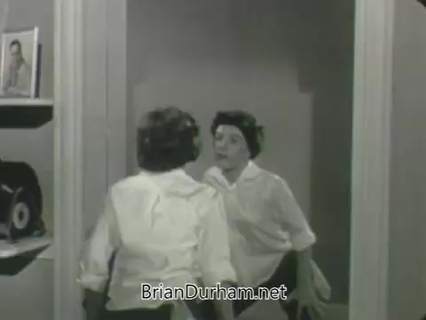 The still from 'ABC Fall 1963 season promos' features a black-and-white scene of a woman looking into a mirror. She appears to be dressed in a casual outfit, with a focused expression as she engages with her reflection. The setting includes some personal items and a photo in the background, suggesting a home environment. Overall, the image conveys a moment of introspection or self-reflection.