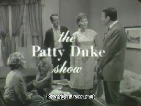 The still features a scene from 'The Patty Duke Show,' showcasing a group of five characters in a domestic setting, with the show's title prominently displayed. The characters appear to be engaged in conversation, suggesting a moment of interaction or conflict. The overall mood reflects a typical sitcom scenario from the early 1960s, characterized by its family-oriented theme.