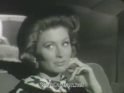 The still features a woman with stylish, voluminous hair and a distinctive patterned outfit, seated in a car. She has a thoughtful expression, lightly resting her chin on her hand, creating an air of sophistication. The background seems to include blurred city scenery, indicative of movement. The overall tone suggests a classic, elegant vibe characteristic of early 1960s television promotions.
