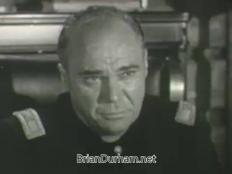 The still features a solemn-looking man in a military uniform, likely a high-ranking officer, seated with a serious expression. He is depicted in close-up, suggesting a moment of tension or contemplation. The black-and-white aesthetic emphasizes the gravity of the scene, typical of mid-20th century promotional content. The setting appears to be an official or command environment.