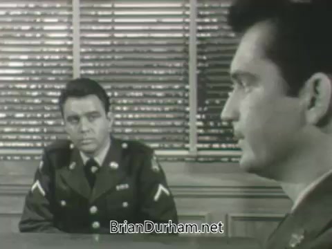The still features two men in military uniforms seated at a table, engaged in a serious conversation. One man, facing slightly away from the camera, appears to be questioning or listening intently, while the other, turned towards the viewer, looks contemplative. The background includes vertical blinds, creating a stark contrast that highlights the tension of their discussion. The scene likely sets the stage for a dramatic moment in the promotional content.