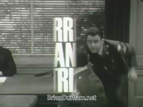 The still features a scene from the ABC Fall 1963 season promos. It shows a man in a military uniform energetically entering a room, appearing to rush or take action. To his left, another man is seated at a desk, likely observing the interaction. Bold, stylized text partially overlays the image, suggesting the show's title or theme, with the letters “RR AN RI” prominently displayed. The setting has a classic mid-20th century office aesthetic, enhanced by the window blinds in the background.