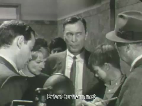 The still features a group of individuals engaged in conversation, set in a hallway. A man in a suit appears to be the focal point, displaying a concerned or serious expression. Surrounding him are several other people, including a woman and a boy, all seemingly focused on the discussion. The scene conveys a sense of intrigue or tension, typical of promotional material from the 1963 ABC fall season.