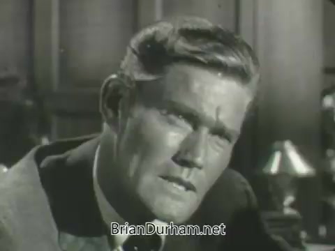 The still features a close-up of a man with a serious expression, possibly in a dramatic or tense moment. He has a well-groomed hairstyle and is dressed in a suit, suggesting a formal setting. The background includes elements like a lamp and what appears to be a bookshelf, indicating a professional or office environment. The overall tone suggests intensity, typical of mid-20th century drama in television promotions.