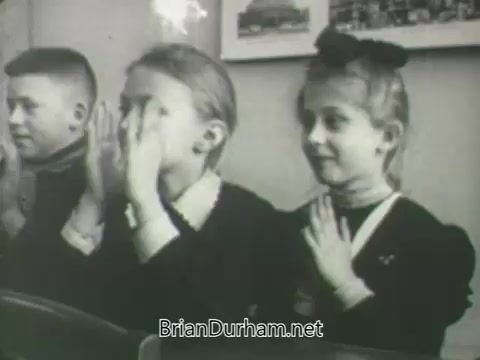 The still from 'ABC Fall 1963 season promos' features a group of children in a classroom setting. Two girls are engaged in a playful activity, with one girl covering her face while the other looks on with a smile. They are dressed in vintage clothing, typical of the early 1960s. The scene captures a nostalgic and innocent moment, emphasizing childhood playfulness and camaraderie.