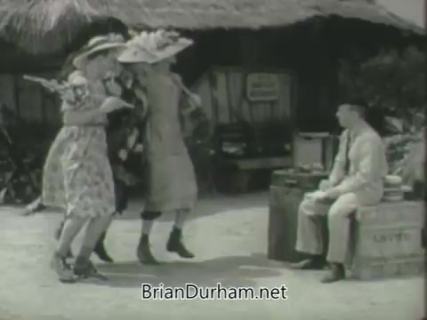 The still features three women in vibrant dresses and large hats energetically dancing or performing in a tropical setting, while a man sits on a crate, seemingly observing them. The background suggests a casual, beach-like atmosphere, typical of mid-20th-century promotional content. The scene exudes a light-hearted, playful vibe, characteristic of television promotions from the era.