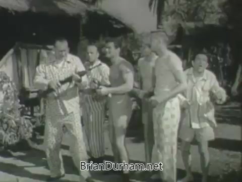 The still from 'ABC Fall 1963 season promos' features a group of six men dressed in casual, tropical-styled clothing, gathered outside a hut. They appear to be engaging in a playful or humorous scene, with one man holding a camera or prop, suggesting a lighthearted or comedic theme. The setting conveys a relaxed, summery atmosphere typical of the era's television promotions.