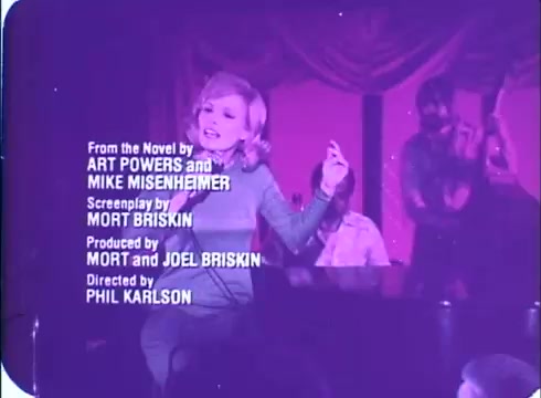 The still from the 'Framed' trailer features a vibrant scene with a woman performing, possibly singing, in a lively atmosphere. She is dressed in a stylish outfit, gesturing expressively. The background hints at a performance setting, likely a nightclub, with warm colors. Text overlays provide credits, mentioning the novel's authors, screenplay, producers, and director. The overall tone conveys energy and entertainment typical of 1970s cinema.