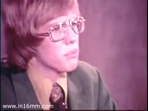 The still from the 'Bicentennial Youth Debates - PSA from 1975' features a young participant, likely preparing for or engaged in a debate. He is wearing a green suit with a collared shirt and patterned tie, indicative of the formal attire typical for the time. The backdrop appears to be a muted color, focusing attention on the subject's expression, which conveys concentration or seriousness. This visual context reflects the program's aim to engage youth in important national discussions during the Bicentennial period, emphasizing civic involvement and critical thinking.