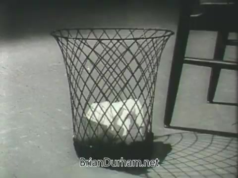 The still from the 'US Army ROTC commercial - 1968' likely features a wire wastebasket, with a block of something white inside. The image has a stark, black-and-white aesthetic, emphasizing simplicity and perhaps conveying a message about responsibility or waste. The presence of a chair nearby suggests a casual setting. The focus on the wastebasket could symbolize choices or consequences, fitting a broader theme of personal accountability in the context of military training.