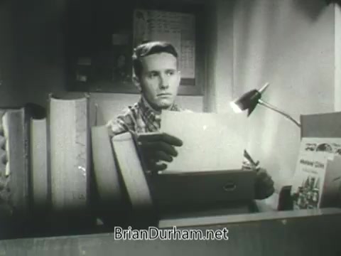 The still depicts a young man sitting at a desk, appearing focused as he reviews a stack of papers. His expression suggests concentration or contemplation. Surrounding him are large books, indicating a study environment, with a desk lamp illuminating his workspace. The black-and-white aesthetic reflects the era of the 1960s, linking it to the context of a US Army ROTC commercial.