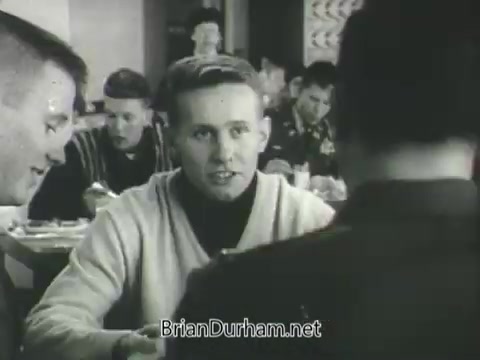 The still from the 1968 US Army ROTC commercial features a gathering, likely in a dining scenario. A young man in a light sweater is engaged in conversation with two others, one of whom is in military uniform. The setting suggests camaraderie and recruitment, highlighting interactions among young men in a military context. The background shows additional individuals, reinforcing a communal atmosphere.
