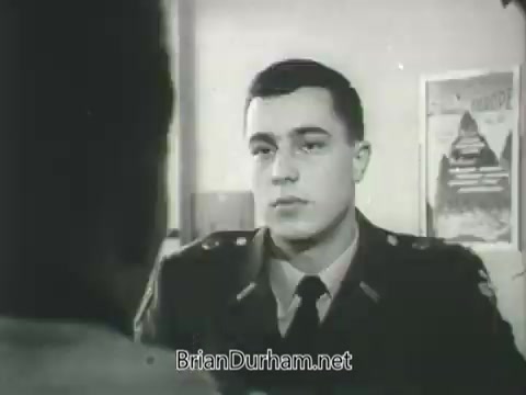The still from the 'US Army ROTC commercial - 1968' features a young man in military uniform, likely an ROTC cadet, engaged in a serious conversation. His expression is focused and attentive, suggesting a discussion about military training or aspirations. The setting appears to be an office, with posters or documents visible in the background, hinting at a formal environment related to military or educational purposes. Overall, the image conveys a sense of ambition and commitment to service.