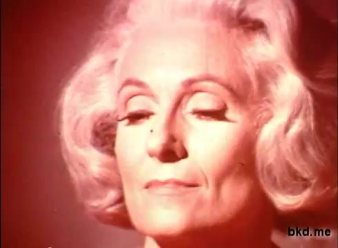 The still features a woman with elegantly styled, silver hair, her eyes closed in a serene expression. The lighting has a warm, reddish hue, creating a soft and inviting ambiance. The focus is on her calm demeanor, suggesting a moment of relaxation, likely associated with the soothing effects of the product being advertised.