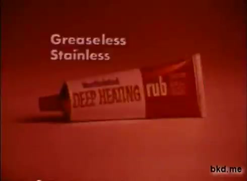 The still features a tube of Mentholatum Deep Heating Rub against a solid, vibrant red background. Prominent text reads 'Greaseless' and 'Stainless,' emphasizing the product's qualities. The tube is angled to showcase its label, highlighting the product name. The overall aesthetic reflects the advertising style of the 1970s, focusing on simplicity and bold colors.