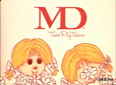 The still from the 1970s 16mm advertisement features a colorful, cartoonish illustration of two girls with blonde hair, each adorned with bows. They have large, expressive eyes and rosy cheeks, holding flowers. Prominently displayed at the top is the 'MD' logo, suggesting it is a promotional piece for MD toilet tissue, highlighted by the tagline 'Twin Play Tissues.' The overall style is playful and nostalgic, characteristic of its era.