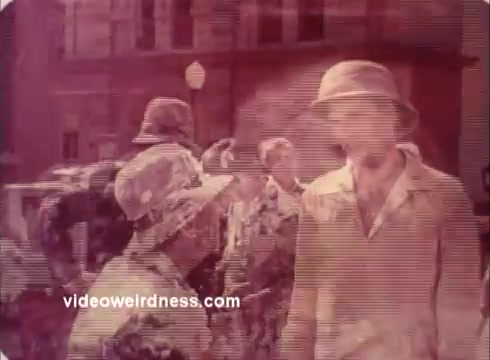 The still from the 'Won Ton Ton' movie trailer features a colorful, slightly faded scene typical of a 16mm print. It shows a group of characters dressed in vintage clothing, including hats. The focus is on two characters engaging with each other, suggesting a comedic or dramatic interaction. The background includes a building and indistinct figures, adding to the bustling atmosphere. The image has a nostalgic quality, indicative of the film's era.