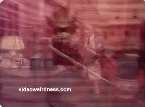 The still from the 'Won Ton Ton' movie trailer features a stylized, faded look typical of 16mm film. It displays a scene filled with various objects and characters, likely showcasing comedic and whimsical elements from the film. The overall color palette leans towards reds and pinks, giving it a vintage feel. The image appears slightly distorted, enhancing its retro aesthetic.