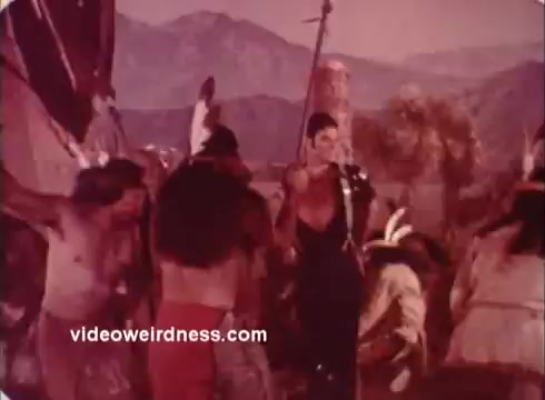 The still from the 'Won Ton Ton' movie trailer depicts a vibrant scene with a central figure dressed elaborately, surrounded by several other individuals in traditional attire. The backdrop features mountainous terrain, contributing to a dramatic atmosphere. The color palette displays warm tones, enhancing the overall vintage feel of the 16mm print. The composition suggests a moment of cultural interaction or ritual.