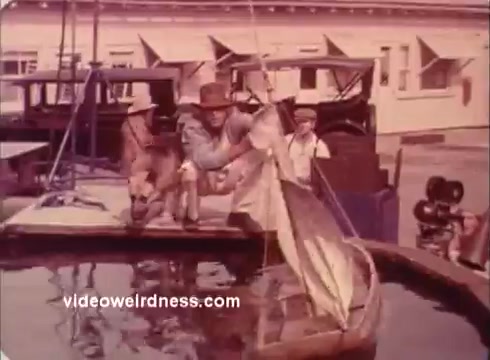 The still from the 'Won Ton Ton' movie trailer features a scene set near a body of water, likely a pond or tank. A man wearing a hat is seen attempting to catch a fish, while two other individuals observe nearby. The setting has a vintage feel, with early 20th-century vehicles in the background, contributing to the film's nostalgic ambiance. The overall tone is lighthearted, hinting at comedic elements typical of the film.