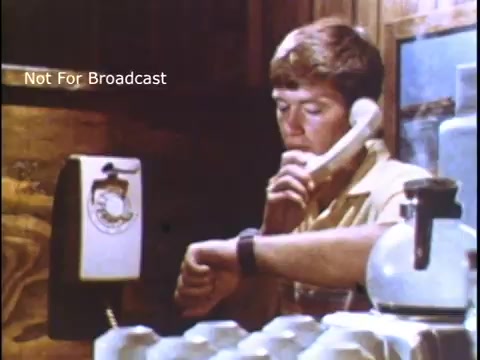 The still depicts a young man in a casual setting, sitting at a wooden table while talking on a rotary phone. He is dressed in a short-sleeve shirt, looking at his watch with a concerned expression. A coffee pot and cups are visible in the foreground, suggesting a domestic space. The scene likely emphasizes the importance of communication and awareness, fitting the PSA context of the US Coast Guard in the 1970s.