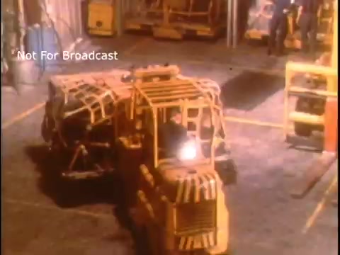 The still features a warehouse environment with industrial equipment, including a yellow forklift and other machinery. Workers are present, engaged in activities typical of a logistics or maintenance operation, likely emphasizing safety procedures. The image conveys a sense of diligence in the context of a public service announcement from the 1970s, focusing on the operations of the US Coast Guard.