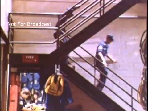 The still shows a scene from a 1970s US Coast Guard PSA, depicting two personnel on a staircase within a building. One individual is descending the stairs, while another is seated nearby, possibly in a relaxed or preparatory stance. The setting appears to be utilitarian, with items such as life jackets visible, emphasizing the operational environment of the Coast Guard. The color tone is characteristic of the period, providing a nostalgic feel.