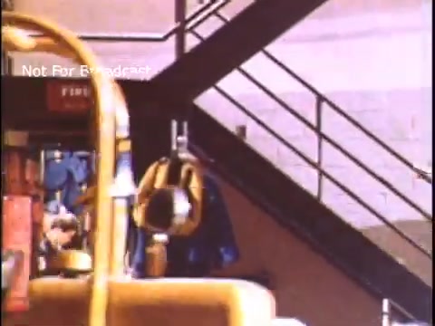 The still from the 1970s US Coast Guard PSA shows a scene inside a facility with stairs in the background. In the foreground, there is yellow equipment, possibly related to rescue operations. The atmosphere suggests a focus on safety and readiness for maritime emergencies. The text 'Not For Broadcast' indicates that it is not intended for public viewing.