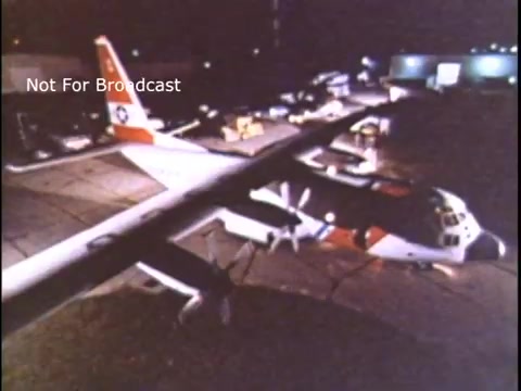 The still features a U.S. Coast Guard aircraft on a tarmac at night, illuminated by artificial light. The plane displays the distinctive red and white color scheme associated with the Coast Guard. In the background, various ground support vehicles and equipment are visible, indicating an active airport setting. The scene conveys readiness and operational activity typical of a public service announcement from the 1970s.