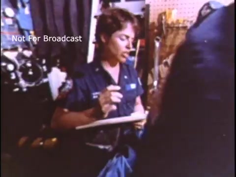 The still shows a female member of the US Coast Guard in a uniform, seated and engaged in taking notes. The background features equipment and tools relevant to the Coast Guard. The retro aesthetic reflects the 1970s style, emphasizing the active role of women in the military during that era.