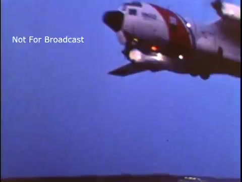 The still features a U.S. Coast Guard aircraft, likely in flight, against a dimly lit sky. The plane is characterized by its distinctive red and white color scheme, emblematic of Coast Guard service. The image conveys a sense of urgency or action, relevant to its purpose as part of a public service announcement from the 1970s.