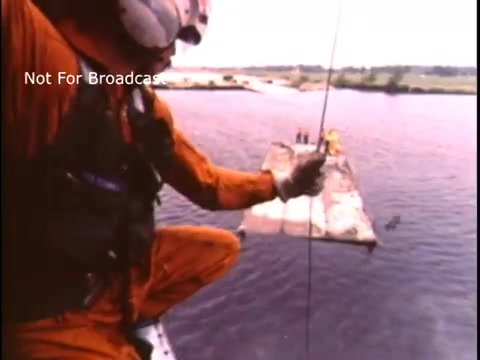 The still shows a US Coast Guard rescuer in an orange uniform, suspended from a helicopter by a rope. He is preparing to assist individuals on a floating platform on the water. The scene captures the urgency and focus of search and rescue operations typical of 1970s PSA campaigns aimed at promoting safety and maritime rescue awareness.