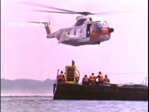 The still depicts a US Coast Guard helicopter hovering above a barge on water, with several individuals in orange life jackets on the barge. The scene suggests a rescue or safety operation, typical of a public service announcement aimed at promoting water safety and the Coast Guard's role in maritime safety during the 1970s. The background features calm water and a pastel sky, creating a focused environment for the ongoing operation.