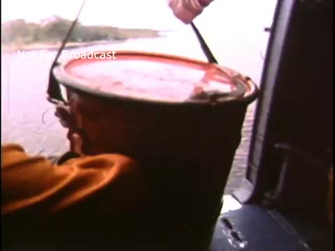 The still shows a person in an orange outfit, likely a Coast Guard member, leaning out of a helicopter. They are holding a red bucket over a body of water, suggesting a rescue or aid operation. The scene captures a dynamic moment in a 1970s public service announcement, emphasizing the Coast Guard's role in safety and rescue efforts.