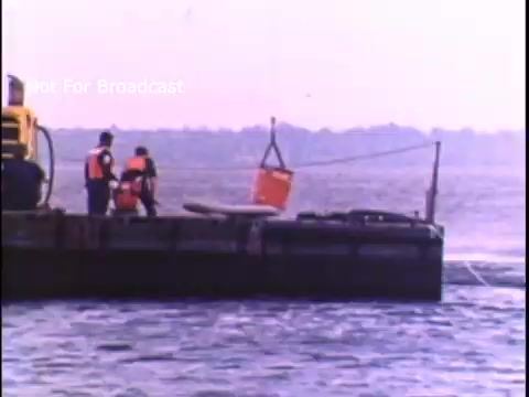 The still depicts two individuals wearing orange life jackets working on a barge in a body of water. They are engaged in lifting a red bucket using a cable, suggesting a demonstration related to safety or rescue operations. The background features a hazy shoreline, indicative of a coastal environment. The overall scene captures an operational aspect of the US Coast Guard from the 1970s.