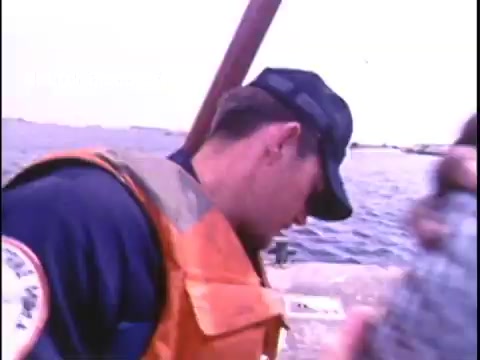 The still depicts a member of the US Coast Guard, dressed in a blue uniform and an orange life jacket, focused on a task near water. The background shows a calm body of water, indicative of a safety or rescue operation. The image captures a sense of duty and vigilance characteristic of Coast Guard missions in the 1970s.