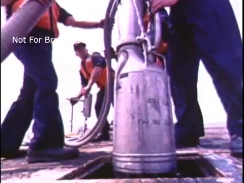 The still features personnel in the U.S. Coast Guard, engaged in a safety procedure on a vessel. They are handling equipment, likely related to fueling or transferring materials, demonstrating operational activities from the 1970s. The focus underscores professionalism and safety protocols within the Coast Guard.