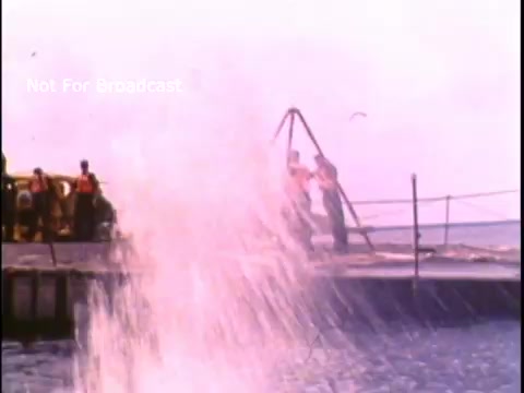 The still from the 1970s US Coast Guard PSA depicts a water scene with a splashing wave, likely from a boat or activity in the water. In the background, several individuals are visible near a pier or dock. The overall aesthetic is characterized by the film grain typical of 16mm footage from that era. The image emphasizes water safety and awareness associated with the Coast Guard's public service announcements.