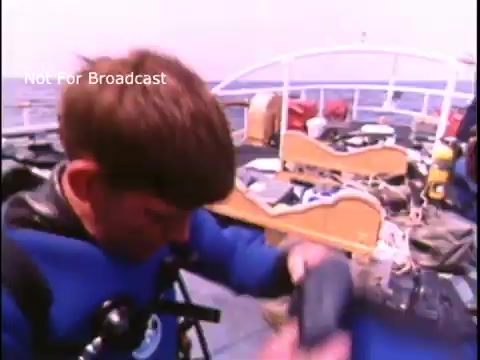 The still depicts a young diver on a boat, preparing for a dive. He is wearing a blue wetsuit and is focused on adjusting his equipment. The background shows various diving gear and the boat’s interior, conveying a maritime safety theme central to the 1970s US Coast Guard public service announcement. The image emphasizes safety and preparedness in diving activities.