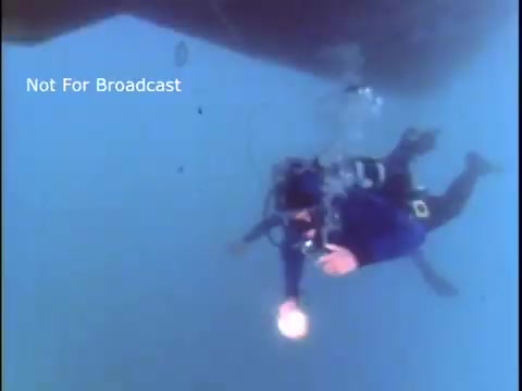 The still features two divers underwater, wearing blue wetsuits and utilizing diving equipment. One diver is holding a bright light source, illuminating the surrounding water. The scene appears to be focused on underwater rescue or safety operations, typical of a US Coast Guard public service announcement from the 1970s. The background includes bubbles and faint visibility, indicative of an underwater environment. The label 'Not For Broadcast' is present, indicating it is a still from a non-broadcast source.
