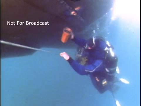 The still depicts a diver in scuba gear underwater, working near the hull of a vessel. The diver holds a container and appears to be securing or retrieving something, suggesting a focus on underwater safety or maintenance. The image is from a 1970s U.S. Coast Guard public service announcement, emphasizing maritime safety and diver operations.