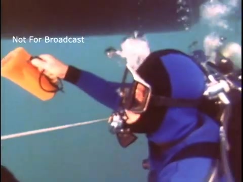 The still features a diver dressed in a blue wetsuit and a diving mask, underwater. The diver is holding an orange object while swimming, indicating an activity related to underwater safety or rescue. Bubbles surround the diver, enhancing the aquatic environment. The image is likely part of a public service announcement by the US Coast Guard from the 1970s, focusing on diving safety or operations.