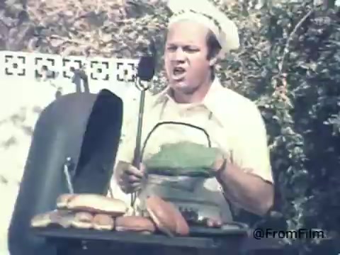 The 16mm still features a man in a chef's hat and a casual shirt, grilling food on a barbecue. His expression is animated, possibly shouting to get attention. The grill has various items, including burgers and hot dogs. The background appears to be a residential outdoor setting, typical of family gatherings or barbecues from the 1970s. This scene likely aims to convey a relaxed, everyday atmosphere, aligning with the promotional theme of the Schick Injector Razor Blades, perhaps highlighting the importance of being well-groomed while enjoying leisure activities.