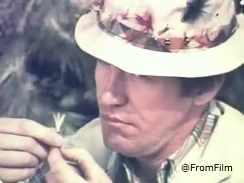 The still from the 1970s commercial for Schick Injector Razor Blades features a man closely examining a razor blade. He is wearing a light-colored hat adorned with feathers and a plaid shirt, suggesting a casual, outdoorsy setting. The focus is on the blade, emphasizing the precision and quality of the product. The overall tone may reflect the era's advertising style, which often combined relatable, everyday scenarios with product demonstrations.