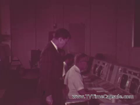 The still from the 1968 American Cancer Society PSA featuring astronaut Walter Schirra captures a moment in a control room setting. Schirra, dressed in a suit, appears to be speaking to individuals seated at control panels, who are likely engaged in monitoring various systems. The environment suggests a serious atmosphere, reflecting the meticulous attention to detail in space missions. Schirra emphasizes the importance of checkups and being alert for warning signs, drawing a parallel between space flight preparations and health vigilance. The overall tone of the PSA is informative, aiming to promote awareness about cancer prevention.