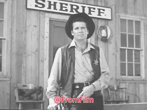 The still from the 1968 Christmas Seals PSA featuring James Garner depicts him in a sheriff's outfit, standing in front of a rustic wooden building marked 'SHERIFF.' He has a serious expression, capturing the essence of his character and the festive yet sincere tone of the announcement. Garner’s demeanor suggests a sense of authority and responsibility, reflecting the message of the PSA during the holiday season.