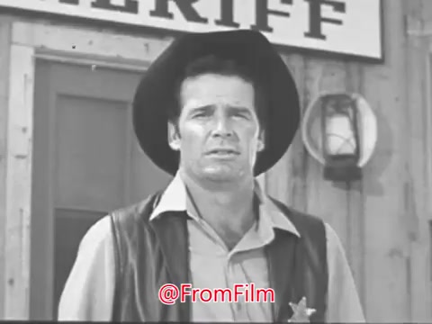 The still from the 'James Garner 1968 Christmas Seals PSA' features James Garner in a setting reminiscent of the Old West. He is dressed in a cowboy outfit with a vest and a sheriff's badge, standing in front of a wooden building with a sign that reads 'SHERIFF.' The scene captures a moment where he addresses the camera, likely sharing a heartfelt message about the holiday season and its significance, as highlighted in the transcript snippet. The overall tone is reminiscent of classic television Westerns, contributing a nostalgic feel to the PSA.
