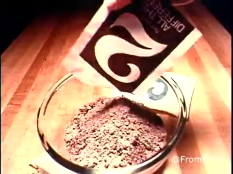 This still from the 1970s Betty Crocker Ring Cake commercial shows a close-up of a hand pouring a packet of powdered mix into a glass bowl. The mix appears to be a dry ingredient, likely for the Bundt cake. The wooden surface beneath the bowl hints at a homey kitchen setting typical of that era. The packaging features a stylized logo, representing the retro aesthetic of the time. Overall, the image captures the essence of home baking and the convenience of using prepared mixes popular during the 1970s.