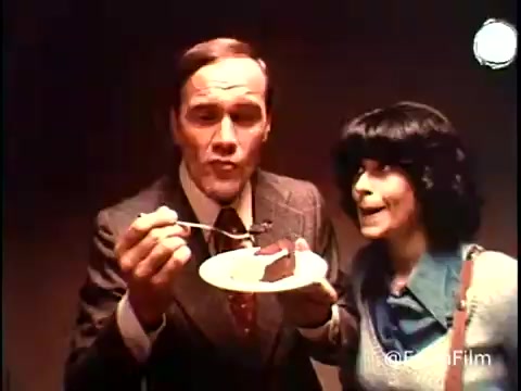 The still from the 1970s Betty Crocker Ring Cake (Bundt cake) commercial features a man in a suit holding a fork with a piece of cake on a plate, appearing to share the cake with a woman beside him. The woman, dressed casually, looks intrigued and excited about the cake. The lighting in the scene is warm, highlighting the cake and emphasizing the enjoyment of the moment. The overall atmosphere conveys a sense of delight and homey charm typical of vintage food commercials.